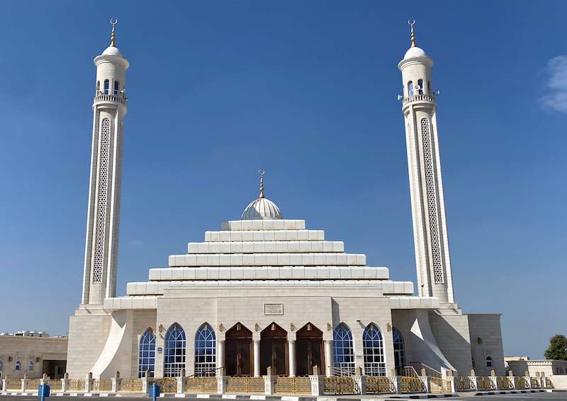 A front view of Ali Mohammed Sadeq Albulushi Mosque with two minarets and a roof resembling a stepped pyramid