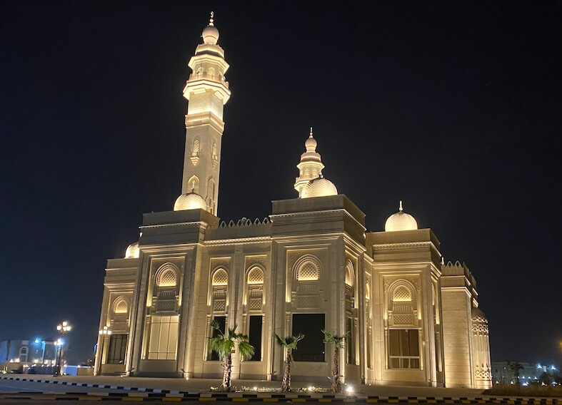 Rear view of large mosque with two minarets, domes lit up at night with black sky background. Image shows the minbar