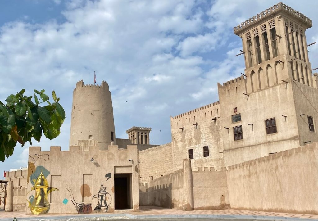 Photo of Ajman Fort and Museum in Ajman, UAE, showcasing traditional Middle Eastern architecture with wind towers and beige stone walls. The foreground features murals of traditional Arabian coffee pots and a leafy tree, all under a partly cloudy sky.