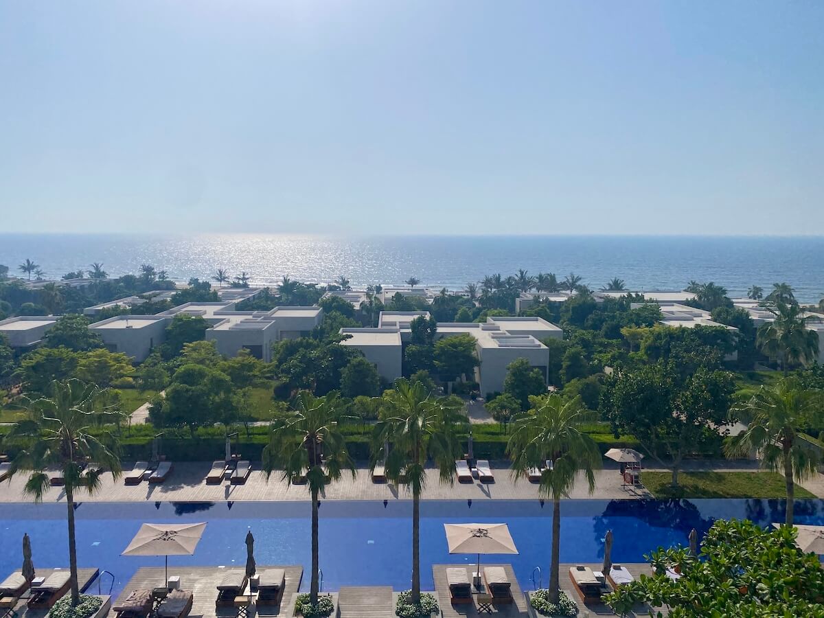 A picturesque view from a balcony at the Ajman Oberoi, featuring a long, blue swimming pool lined with sun loungers and umbrellas. Beyond the pool, lush greenery and modern villas stretch towards the sparkling sea under a clear blue sky.