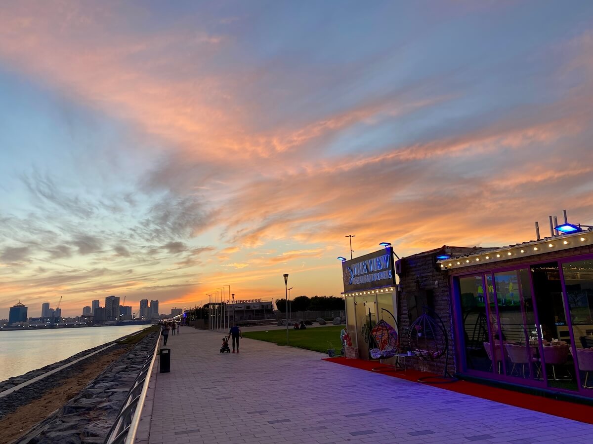 Al Zorah Marina in Ajman, UAE, at sunset, with a waterfront walkway, a brightly lit restaurant, and the city skyline in the distance. The sky is painted with vibrant hues of orange, pink, and blue.