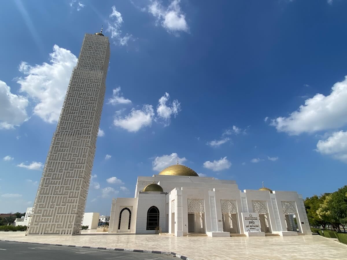 Amina bint Al Ghurair Mosque in Ajman, UAE, featuring a tall minaret with Arabic calligraphy covering it and a gold-domed main building against a bright blue sky with scattered clouds. The mosque has a modern architectural design.