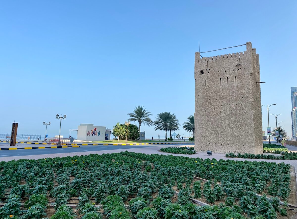 Murabba Tower in Ajman, UAE, with a traditional stone structure surrounded by green plants and palm trees. A "I ♥ Ajman" sign is visible in the background, along with a clear blue sky.