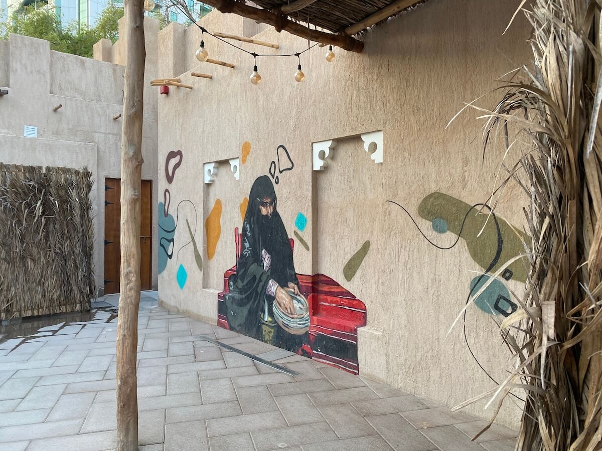 A colourful mural in the Ajman heritage district depicting a woman in traditional attire, sitting and working with her hands on a flat surface. The surrounding area features natural, earthy textures and materials, enhancing the rustic and cultural ambiance.