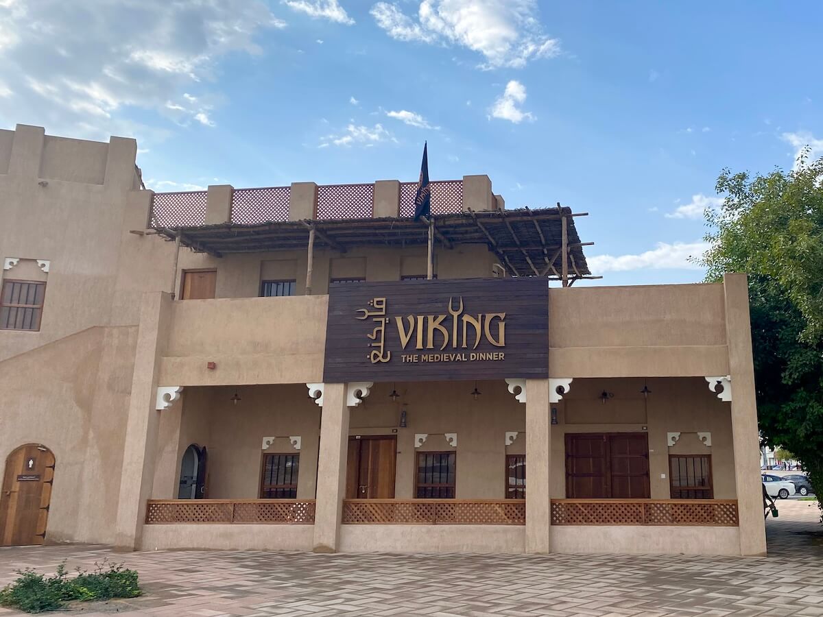 The front of The Viking Medieval Diner in Ajman, featuring a light brown building with wooden accents and a sign displaying the restaurant's name. The exterior includes traditional architectural elements and a small balcony, with a partly cloudy sky in the background.