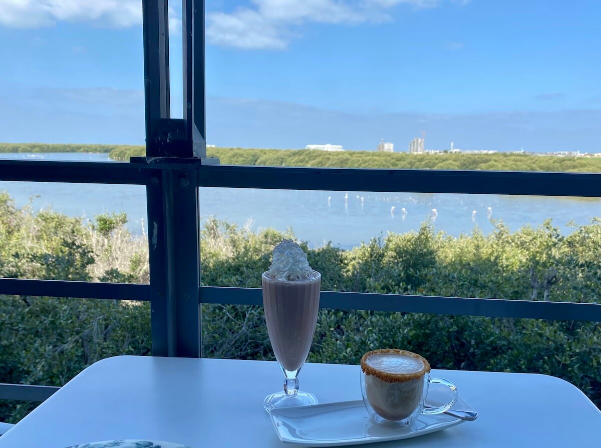 A scenic view from the Al Zorah Golf Pavilion terrace, showing a milkshake topped with whipped cream and a cappuccino on a white table. In the background, a serene landscape with a lush mangrove forest, a waterway with flamingoes, and distant buildings under a bright blue sky.