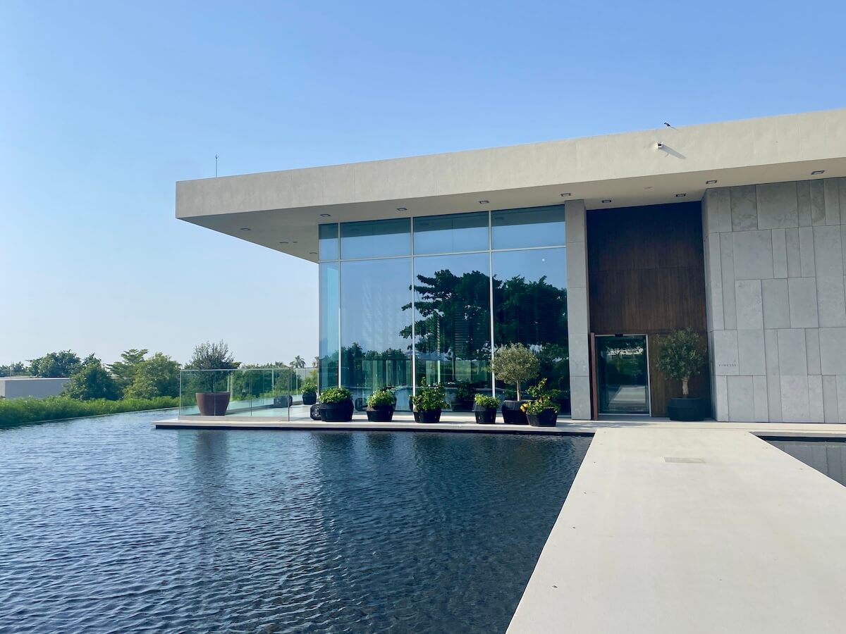 A modern building with large glass windows and a flat roof, surrounded by a reflective water feature and potted plants. The clear blue sky and greenery in the background add to the serene atmosphere. This elegant structure is part of the Vinesse Restaurant at the Oberoi Al Zorah in Ajman.