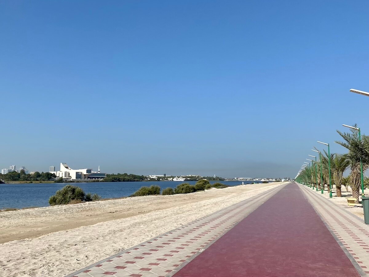 A view of the Al Qurm walking track in Ajman, featuring a long, red path flanked by patterned tiles, lined with palm trees and solar lights. To the left, there's a sandy beach and a waterway, with buildings and mangroves in the distance under a clear blue sky.