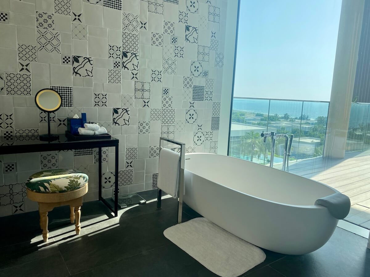 A modern bathroom featuring a freestanding white bathtub positioned near a large window with a scenic ocean view. The wall is decorated with a variety of patterned tiles, and there is a vanity table with a mirror and a cushioned stool. The setting is part of a luxurious suite at the Oberoi Al Zorah in Ajman.