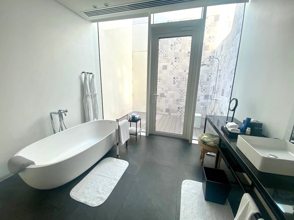 A contemporary bathroom with a freestanding bathtub, a vanity with a mirror and stool, and large windows allowing natural light. The bathroom leads to an outdoor shower area adorned with decorative tiles. Two white robes hang next to the tub, adding to the luxurious ambiance of this villa at the Oberoi Al Zorah in Ajman.