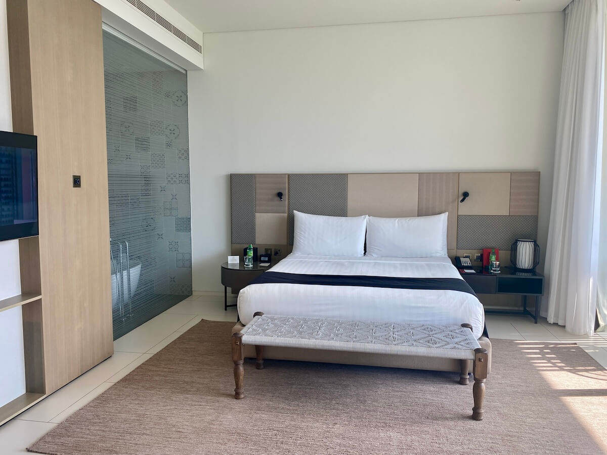 A modern hotel room with a large bed featuring white linens and a black runner, a padded headboard, and a bench at the foot of the bed. The room includes a glass-enclosed bathroom with patterned tiles and natural light streaming in through floor-to-ceiling windows. This elegant space is a Premier Ocean View Room at the Oberoi Al Zorah in Ajman.