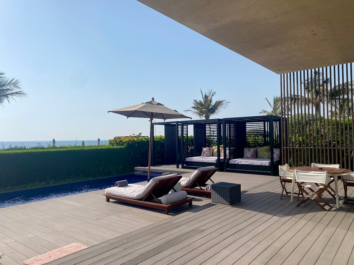 A spacious outdoor deck featuring a private pool, sun loungers with white cushions under umbrellas, and shaded cabanas with plush seating. The area includes a dining table and chairs, surrounded by lush greenery and palm trees, with a view of the ocean in the background. This luxurious setup is part of a villa at the Oberoi Al Zorah in Ajman.